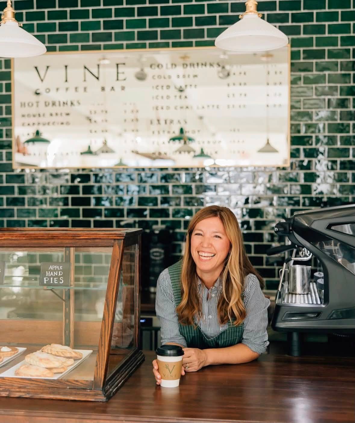 Photo of a smiling woman with a coffee in her hand behind the barista bar.